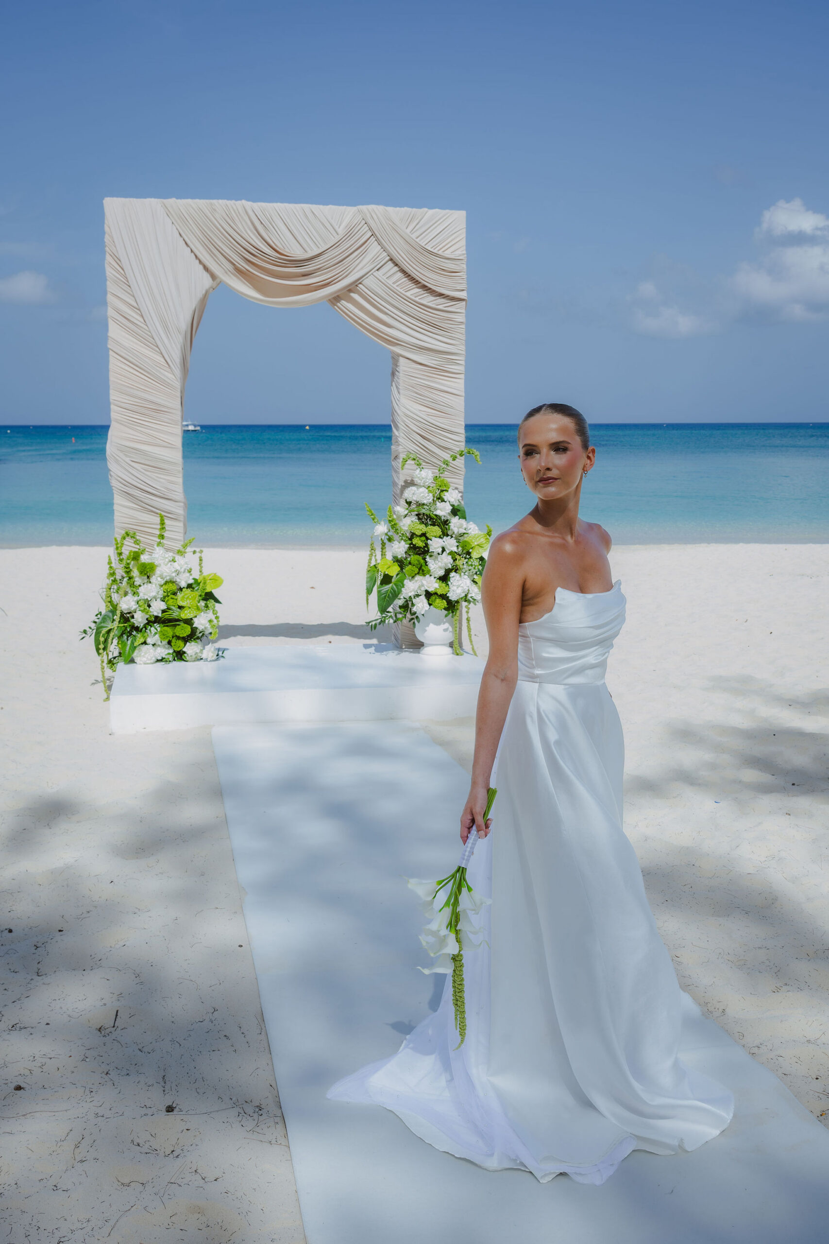 Bride in a white wedding gown holding a bouquet beneath a floral ceremony arch on the beach at The Westin Grand Cayman.